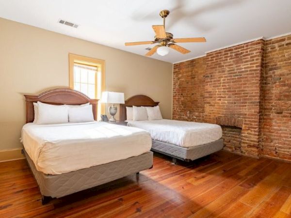 The image shows a bedroom with two beds, brick accent wall, wooden floor, ceiling fan, and a window letting in natural light.