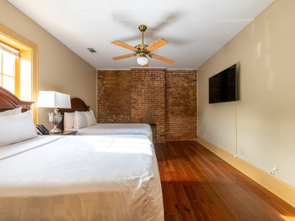 The image shows a bedroom with two beds, a brick accent wall, ceiling fan, and flat-screen TV, all bathed in natural light from a window.