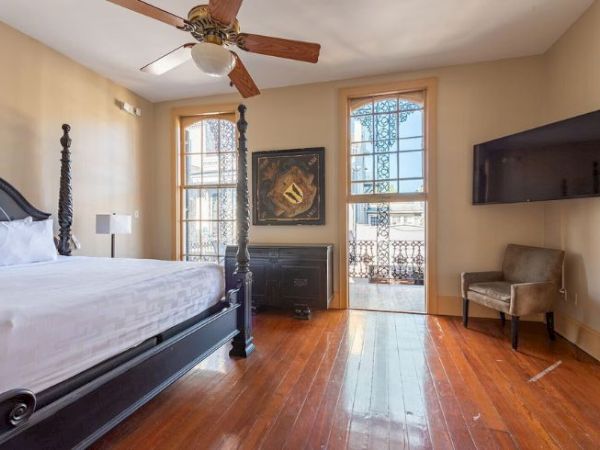 The image shows a bedroom with a large bed, ceiling fan, TV, wooden floor, and windows letting in natural light, creating a cozy atmosphere.