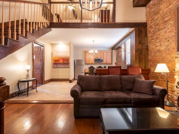 The image shows a cozy living room with a brown sofa, brick wall, staircase, and open kitchen. Warm lighting adds to the inviting atmosphere.