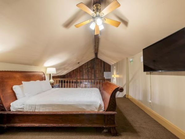 A bedroom with a large wooden bed, ceiling fan, lamp, and wall-mounted TV, located in a loft space with carpeting and sloped ceiling.