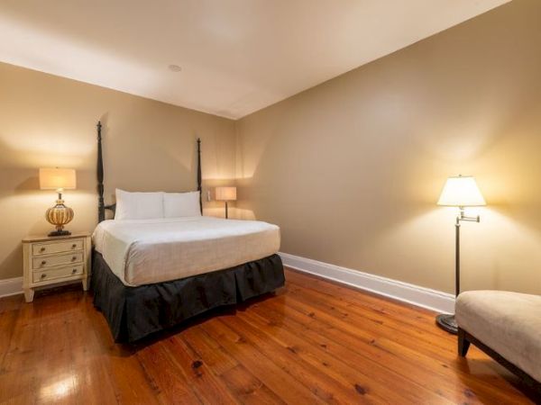 A cozy bedroom with a bed, two bedside lamps, a chair, and hardwood flooring. Neutral wall color adds warmth and simplicity.