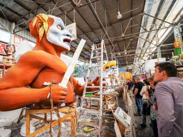 A sculptor builds a large orange figure wearing a white mask in a busy workshop, with ladders, tools, and onlookers nearby, all under a high ceiling.