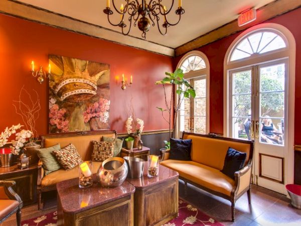 Cozy lounge with warm red walls, a chandelier, leather couch, rustic coffee table, potted plants, art, and a glass-paned door letting in natural light.
