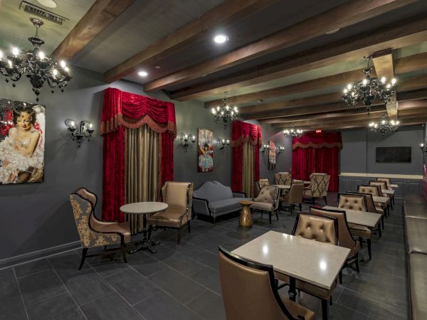 A stylish, dimly lit lounge with red curtains, chandeliers, and cream chairs around square tables, creating an intimate cafe vibe.