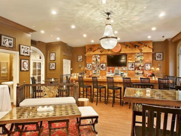 A cozy dining room or cafe with warm wood tones, framed photos on the walls, a central chandelier, and several tables and chairs neatly arranged, plus a TV on the back wall.