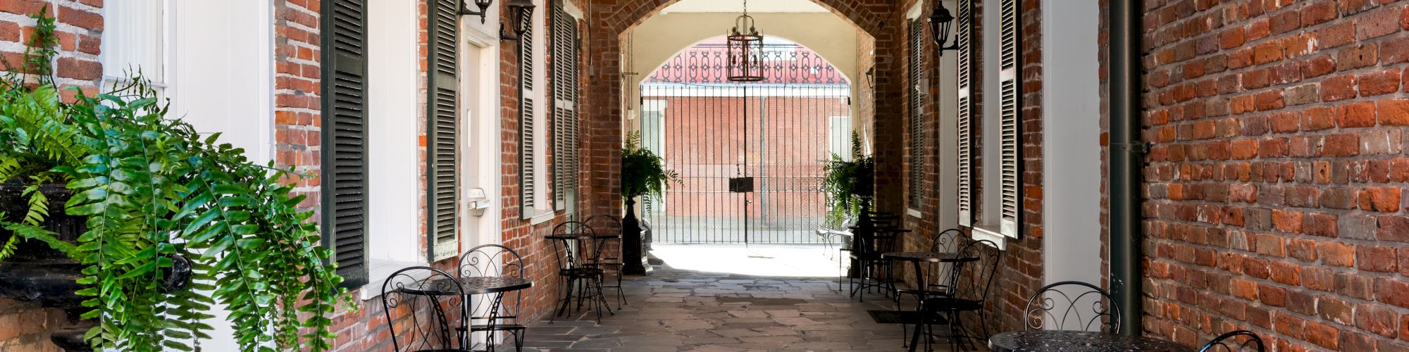 A brick-pathed alley between buildings with small black tables and chairs, potted greenery, arches, and lanterns&mdash;cozy outdoor caf&eacute; vibe.