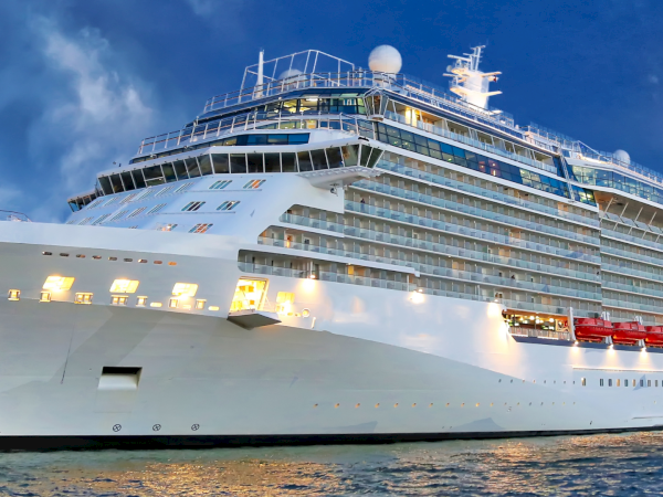 A massive white cruise ship docked at sunset, multiple decks full of windows and balconies, with bright lights reflecting on calm water.