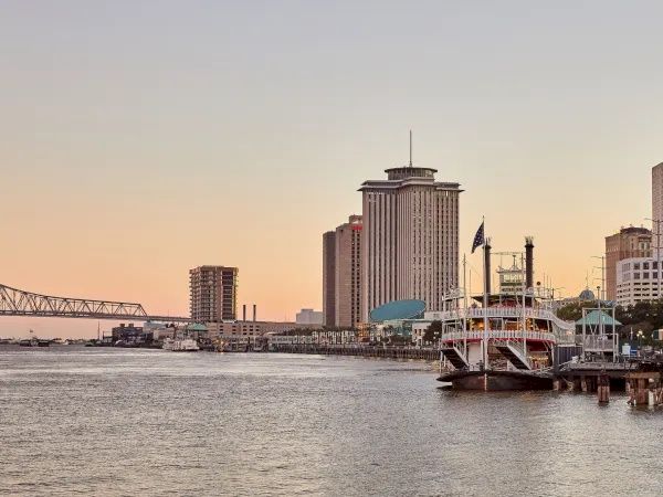 A riverside city skyline at sunset with a tall tower and a docked ferry/ship by the pier.