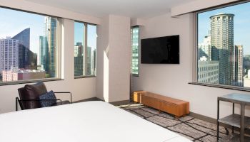 A modern hotel room with city views, a bed, wall-mounted TV, armchair, and a small table with windows overlooking skyscrapers.