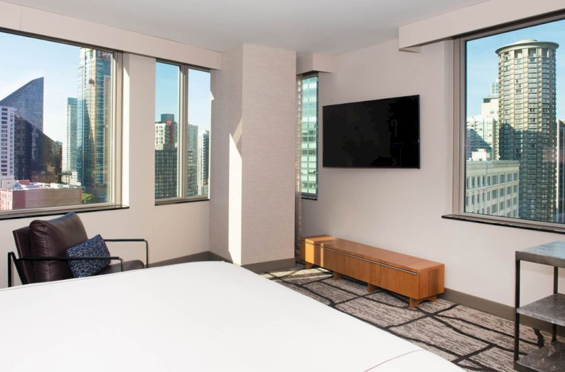 A modern hotel room with city views, a bed, wall-mounted TV, armchair, and a small table with windows overlooking skyscrapers.