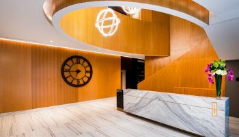 A modern lobby features a marble desk, wall clock, spiral wooden decor, and hanging lights, with flowers atop the counter.