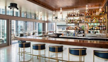 This image shows a modern bar with high chairs, a stocked shelf of bottles, and large windows offering a city view.