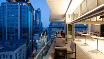 A modern rooftop terrace overlooks a cityscape with tall buildings, featuring seating and open, illuminated indoor space to the right.
