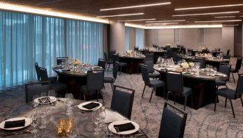 A banquet hall with round tables set with black napkins, glassware, and white flowers, featuring modern lighting and carpeted floors.