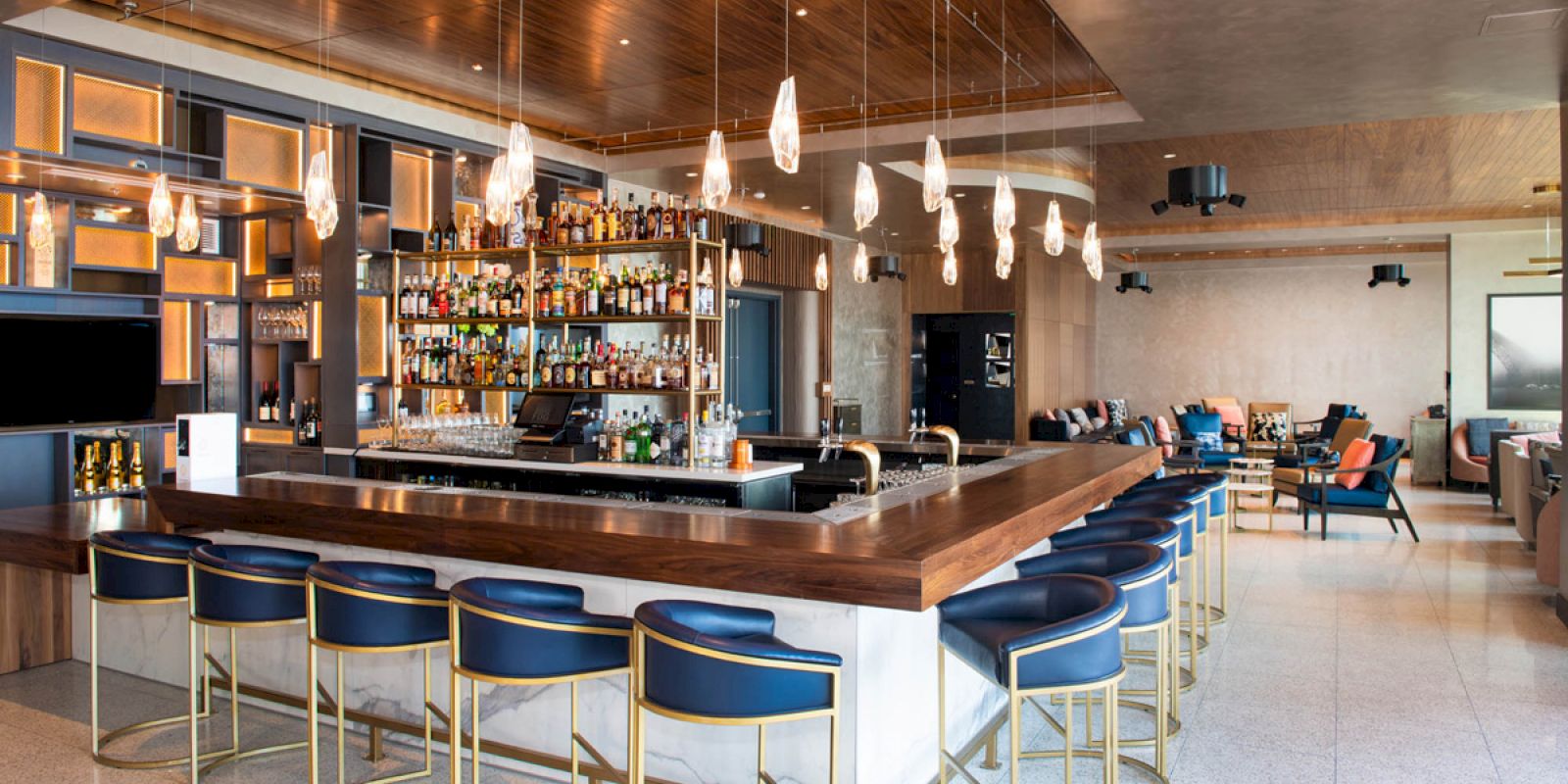 A modern bar with a central counter, blue upholstered stools, hanging lights, and a variety of bottles on display behind the counter.