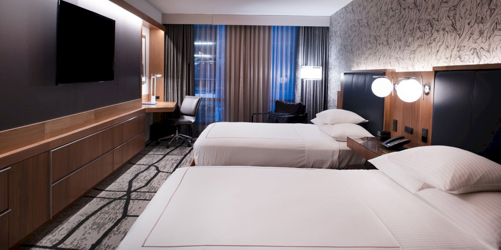 The image shows a modern hotel room with two beds, a flat-screen TV, a desk, and a patterned carpet, creating a cozy atmosphere.