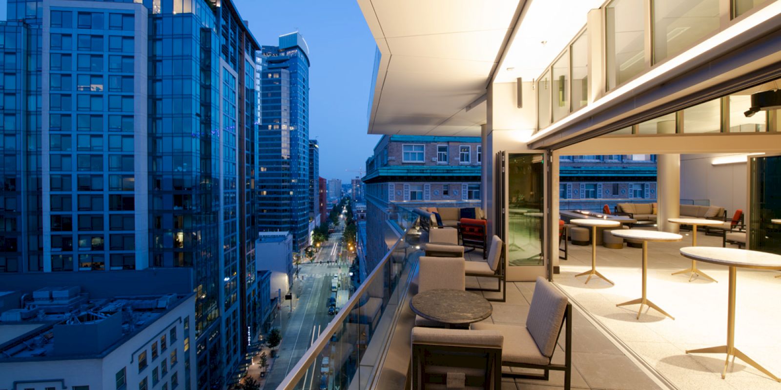 A modern rooftop terrace overlooks a city street lined with tall buildings, featuring seating and tables under ambient evening lighting.