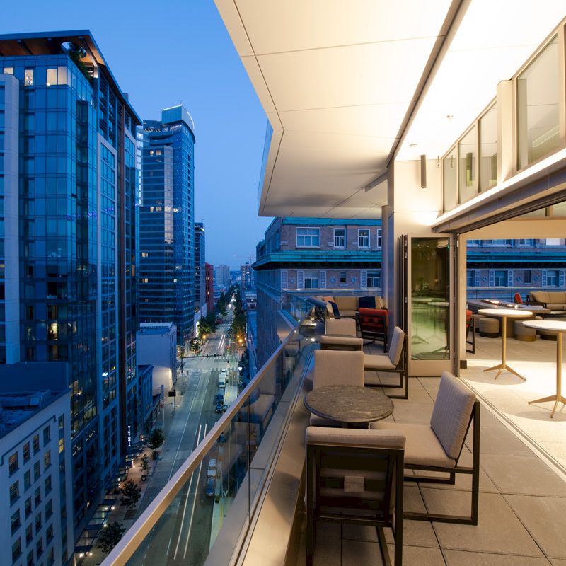 A modern balcony view at twilight with city skyscrapers, outdoor seating, and warm interior lighting, creating an inviting ambiance.