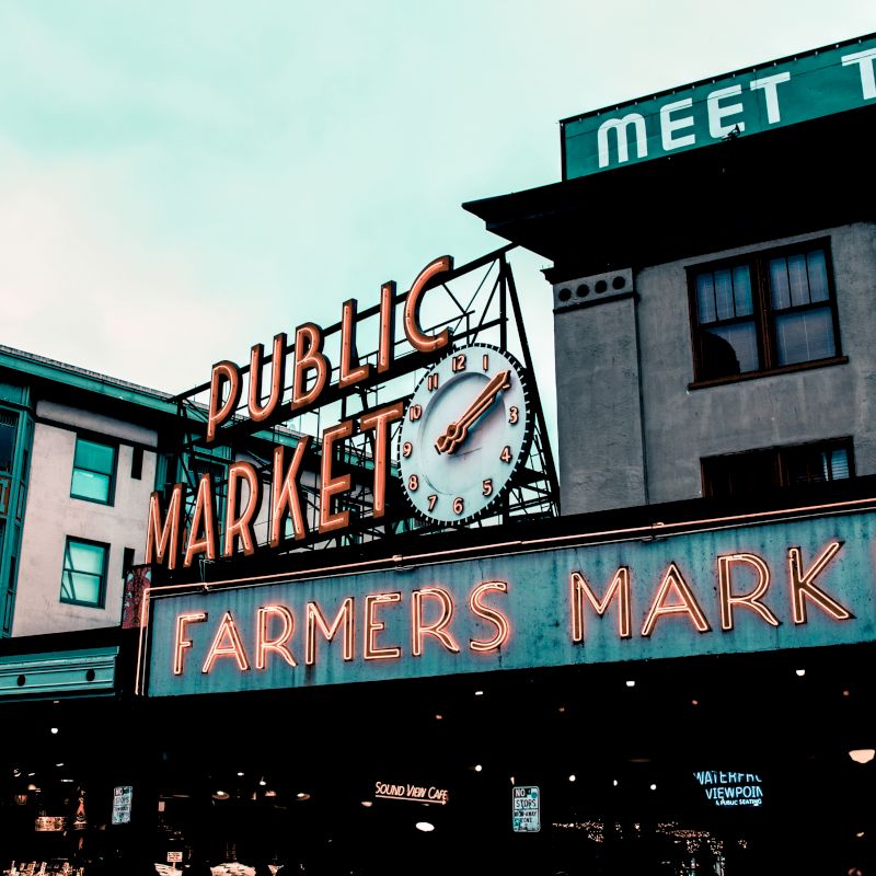 The image shows a neon sign reading "Public Market" and "Farmers Market" with a clock, set against urban buildings under an overcast sky.