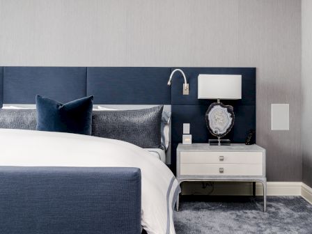 A modern bedroom with a blue headboard, white bedding, navy throw pillows, and a sleek nightstand with a lamp and decorative item.