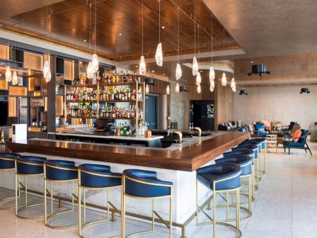A modern bar with blue stools, a well-stocked liquor shelf, pendant lighting, and lounge seating in a sleek, stylish interior setting.