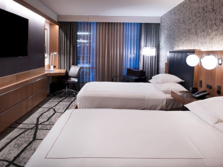 A modern hotel room with two beds, a desk, a TV on the wall, and a patterned carpet, all in a minimalist design.