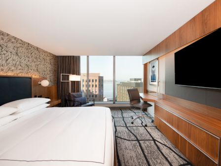 A modern hotel room with a large bed, wall-mounted TV, desk, chair, and a window view of city buildings and the water.