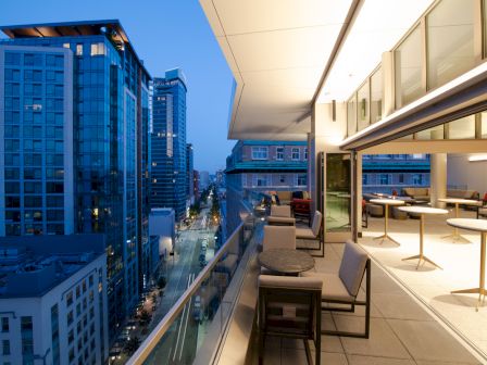 A modern cityscape viewed from an elevated balcony at dusk, featuring outdoor seating and a lit indoor area visible through large windows.