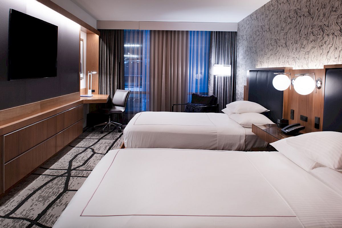 The image shows a modern hotel room with two beds, a TV, a desk, and stylish decor.