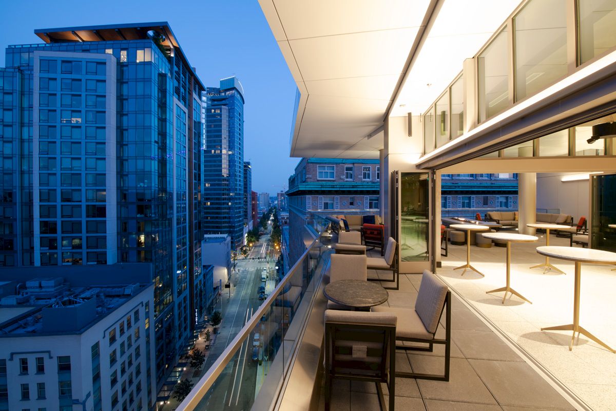 A modern balcony view shows tall buildings and a street below at dusk, featuring seating and lit interior space.