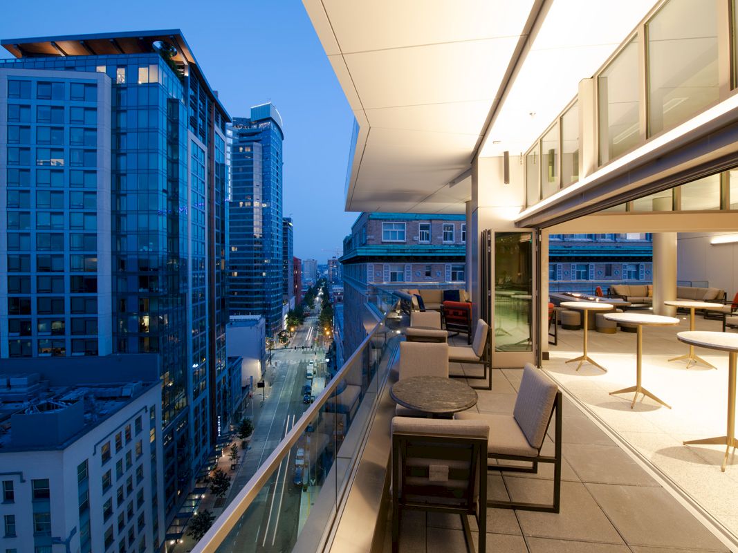 A modern balcony view shows tall buildings and a street below at dusk, featuring seating and lit interior space.