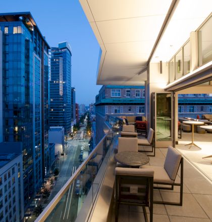 A modern cityscape with tall buildings at dusk, featuring a sleek balcony with seating and tables overlooking the street below.