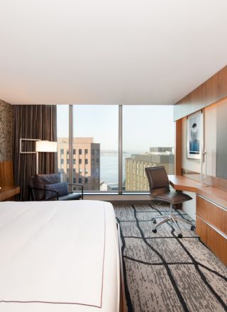 A modern hotel room features a large bed, wall-mounted TV, desk, chair, large windows, and stylish decor.