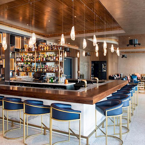 The image shows a modern bar with stylish seating, illuminated bottle shelves, and pendant lights, creating a sleek and inviting atmosphere.