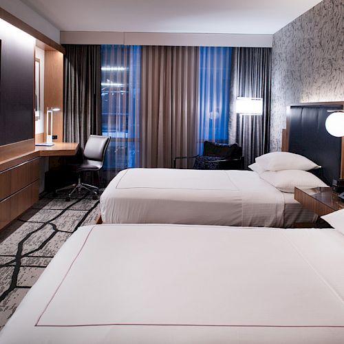 The image shows a modern hotel room with two beds, a flat-screen TV, a desk with a chair, and stylish lighting and decor.