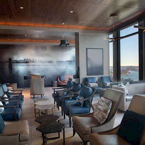 A modern lounge with plush seating, large windows offering a waterfront view, and a wall mural depicting a city skyline in the background.