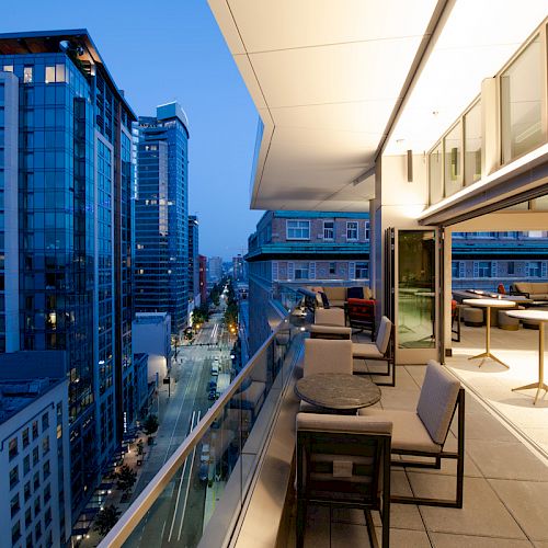 A modern rooftop patio with tables and chairs overlooking a city street, surrounded by tall buildings at twilight.