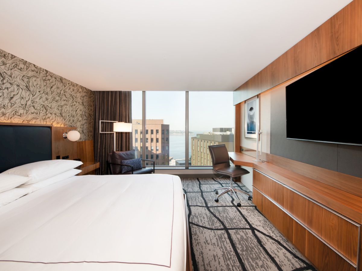 A modern hotel room with a large bed, desk, and chair, featuring cityscape views through large windows, wood paneling, and a large TV.