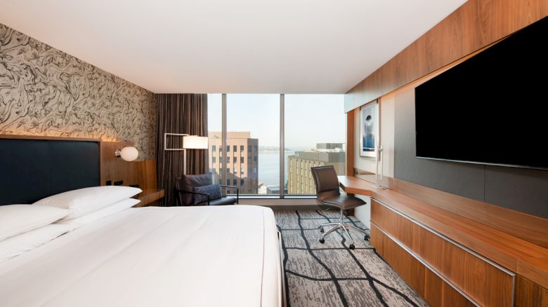 A modern hotel room with a large bed, desk, and chair, featuring cityscape views through large windows, wood paneling, and a large TV.