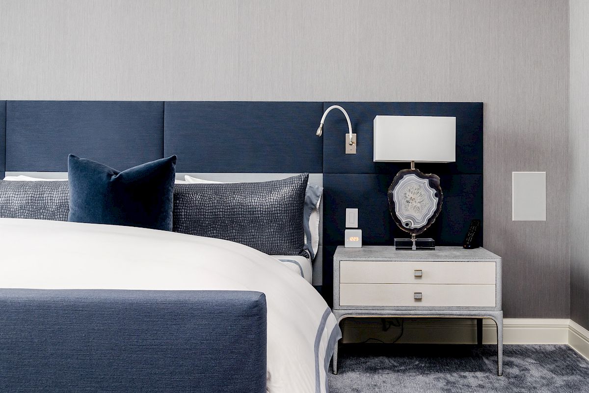 A modern bedroom features a navy headboard, white bedding, a nightstand with a lamp, and a decorative item, creating a sleek, minimalist look.