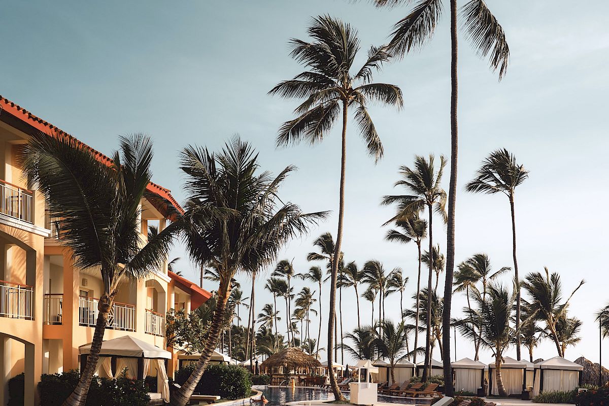 A tranquil poolside scene with lounge chairs, palm trees, and a hotel building under a clear sky, creating a relaxing atmosphere.