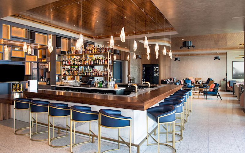The image shows a stylish bar with blue stools, wooden counters, pendant lights, and a well-stocked back bar, set in a modern lounge area.