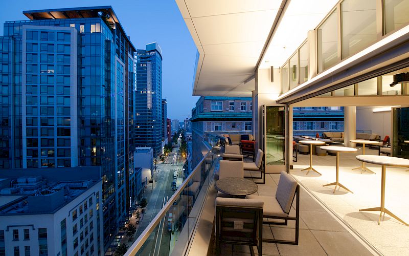 A modern rooftop terrace overlooking a cityscape at dusk, featuring outdoor seating, glass railings, and illuminated interiors.