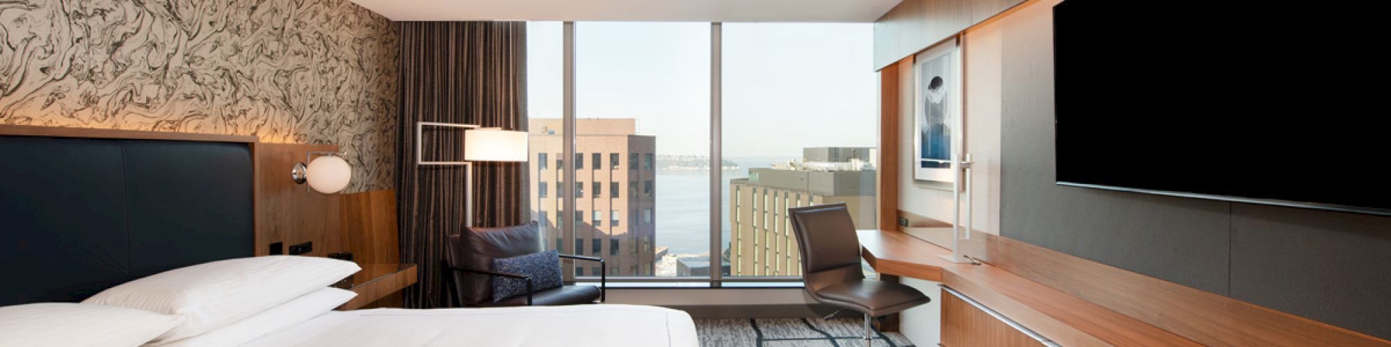 A modern hotel room features a large bed, flat-screen TV, desk, and chair with a city view through a large window.